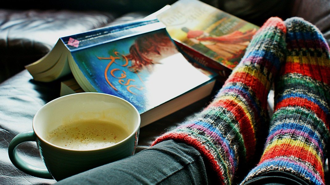 Zdjęcie kolorowe. Na nim stopy ubrane w kolorowe skarpety, filiżanka kawy i rozłożona książka okładką do góry.