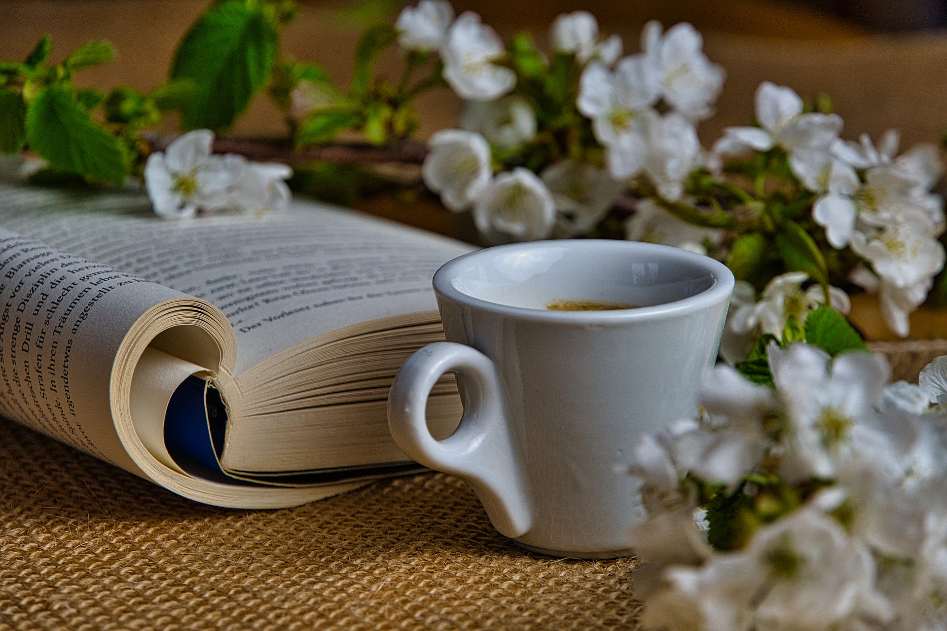 Zdjęcie kolorowe, na stoliku otwarta książka, kubek z kawą obok kwitnąca gałązka